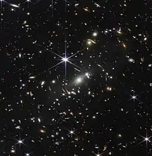 The background of space is black. Thousands of galaxies appear all across the view. Their shapes and colors vary. Some are various shades of orange, others are white. Most stars appear blue, and are sometimes as large as more distant galaxies that appear next to them. A very bright star is just above and left of center. It has eight long, bright-blue diffraction spikes. Between 4 o'clock and 6 o'clock in its spikes are several very bright galaxies. A group of three are in the middle, and two are closer to 4 o'clock. These galaxies are part of the galaxy cluster SMACS 0723, and they are warping the appearances of galaxies seen around them. Long orange arcs appear at left and right toward the center.